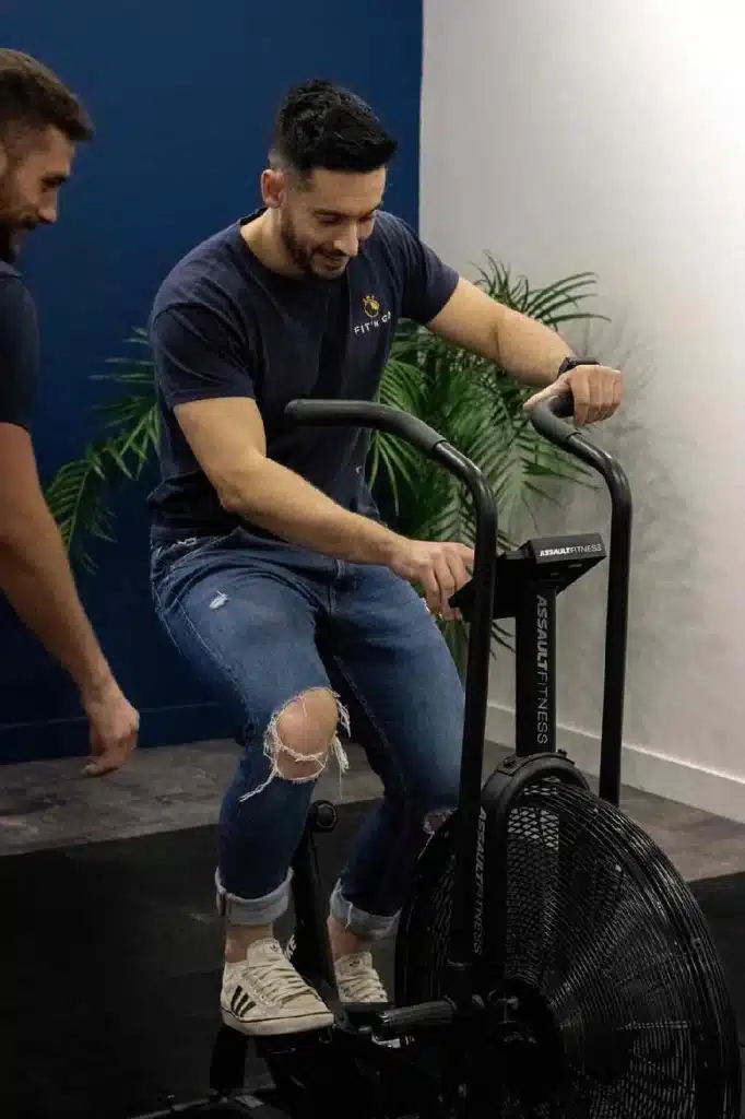 Homme souriant s'entraînant sur un vélo Assault Fitness en jean et t-shirt bleu dans une salle de fitness.