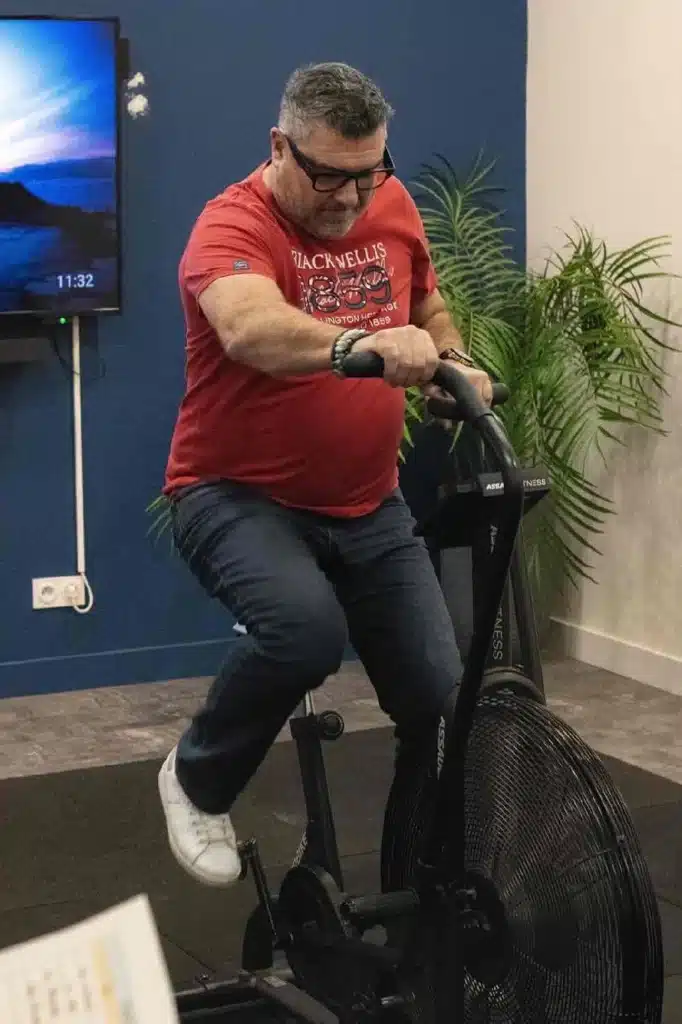 Homme en t-shirt rouge Blackwellis pédalant sur un vélo à air Assault Fitness dans une salle de sport.