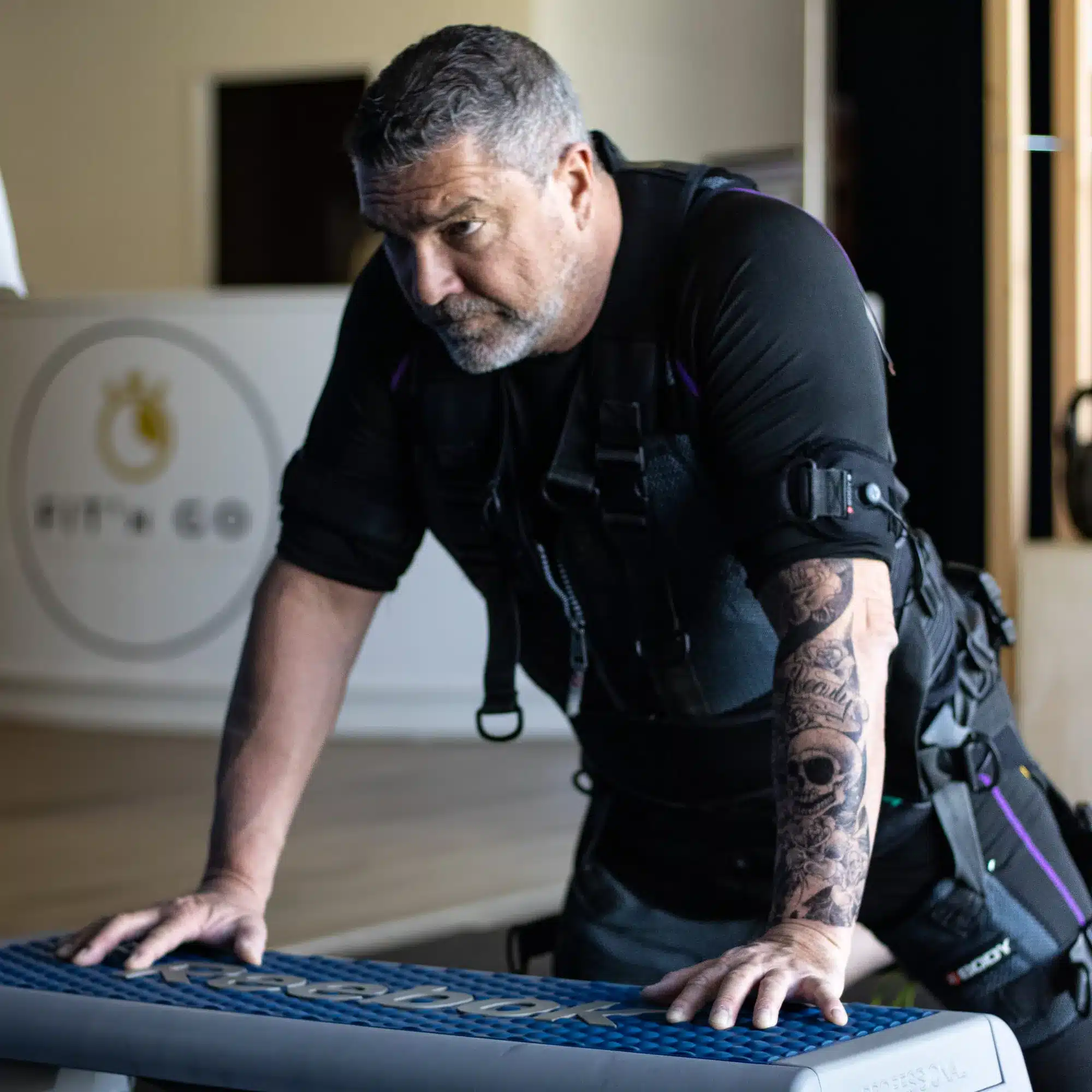 Homme tatoué en séance d'EMS training Fit'n Go, mains sur un step Reebok, exprimant l'effort physique.