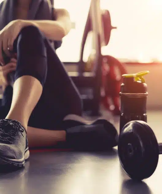Séance de Musculation : Repos et Hydratation en Salle Femme se reposant après une séance de fitness avec gourde, haltère et baskets de sport dans une salle de musculation.